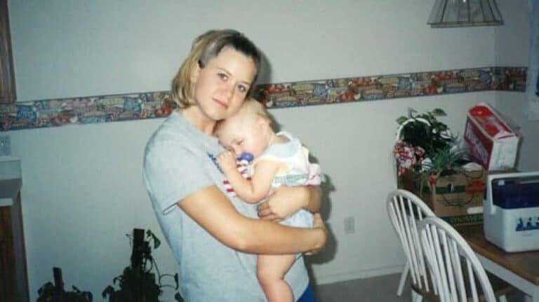 A woman holding a sleeping baby in a kitchen with a table, chairs, and a potted plant in the background.