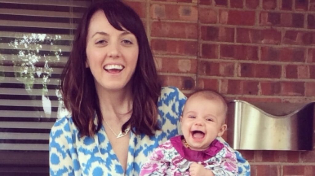 Woman in a blue and white patterned shirt holding a smiling baby outside in front of a brick wall. Both are smiling.