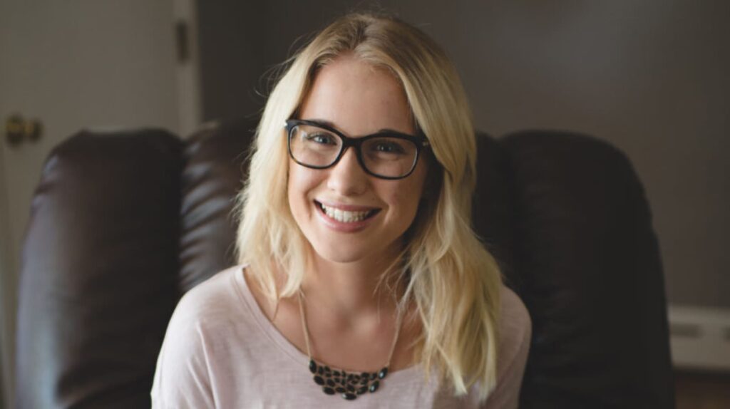 A blond woman wearing glasses and a purple top smiles while sitting on a dark brown leather chair indoors.