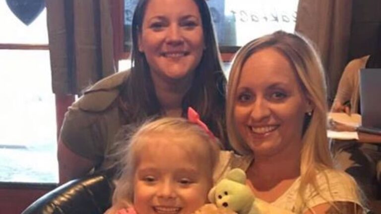 Two women smile at the camera while one holds a young girl with a bow in her hair and a stuffed toy. They are indoors with natural light.