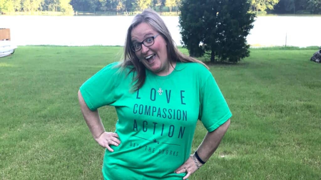 A smiling woman wearing glasses and a green T-shirt with the words "LOVE COMPASSION ACTION" poses with hands on hips in a grassy area.