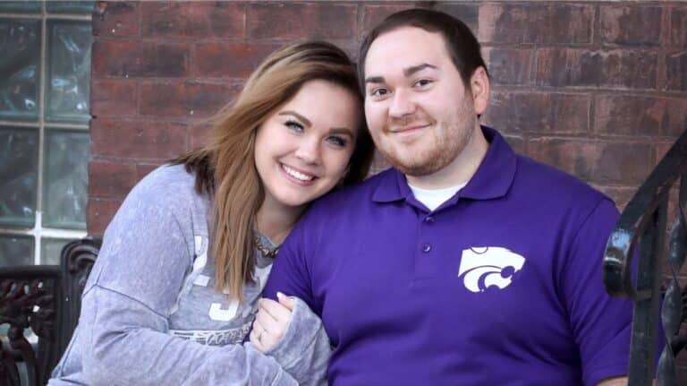 A smiling woman leans on a smiling man. The man wears a purple shirt with a white logo, and they sit against a brick wall.