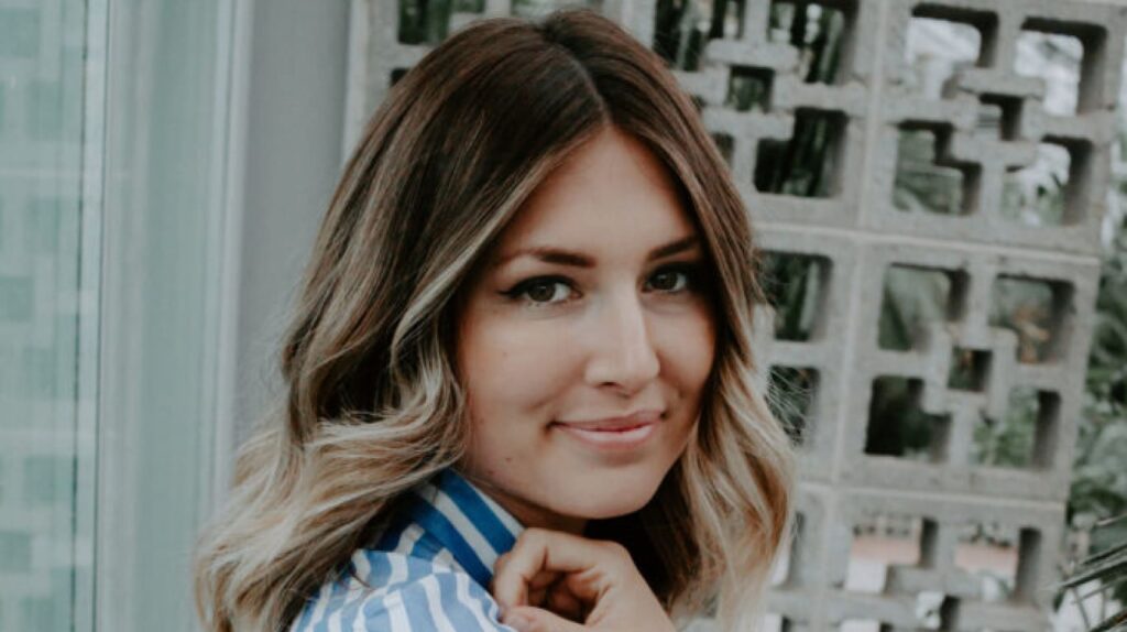 A woman with wavy brown hair and blonde highlights, wearing a striped shirt, smiles while looking at the camera with a patterned wall behind her.