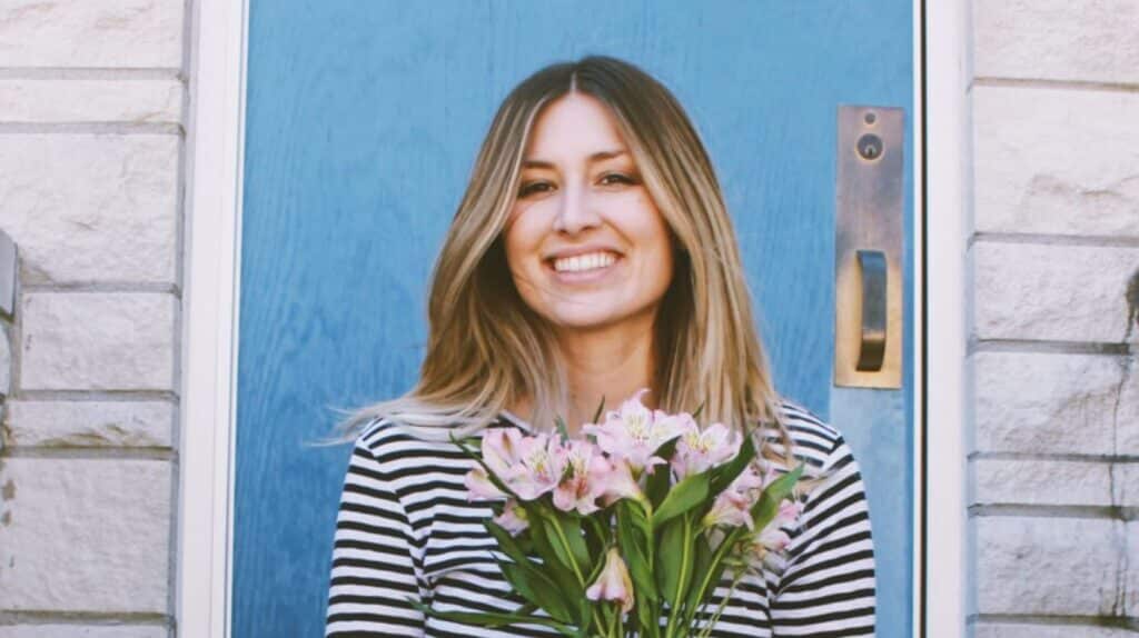 Smiling woman with long hair stands in front of a blue door, holding a bouquet of pink flowers, wearing a black and white striped shirt.