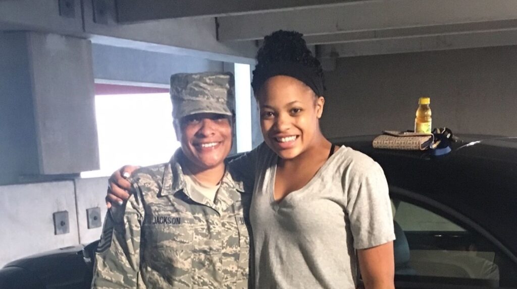 Two individuals, one in a military uniform and one in casual attire, smiling and standing close together in a parking garage.