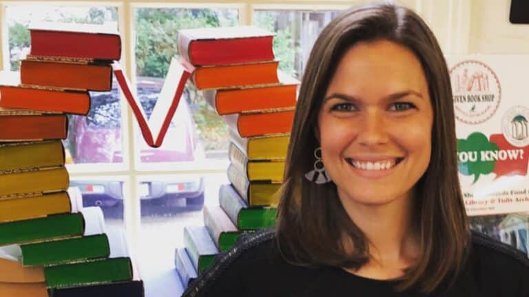 A smiling woman stands in front of a V-shaped arrangement of colorful books, with a window in the background.