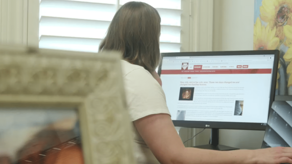 A person sitting at a desk, facing a computer monitor displaying a webpage with text and images, next to a framed photo.