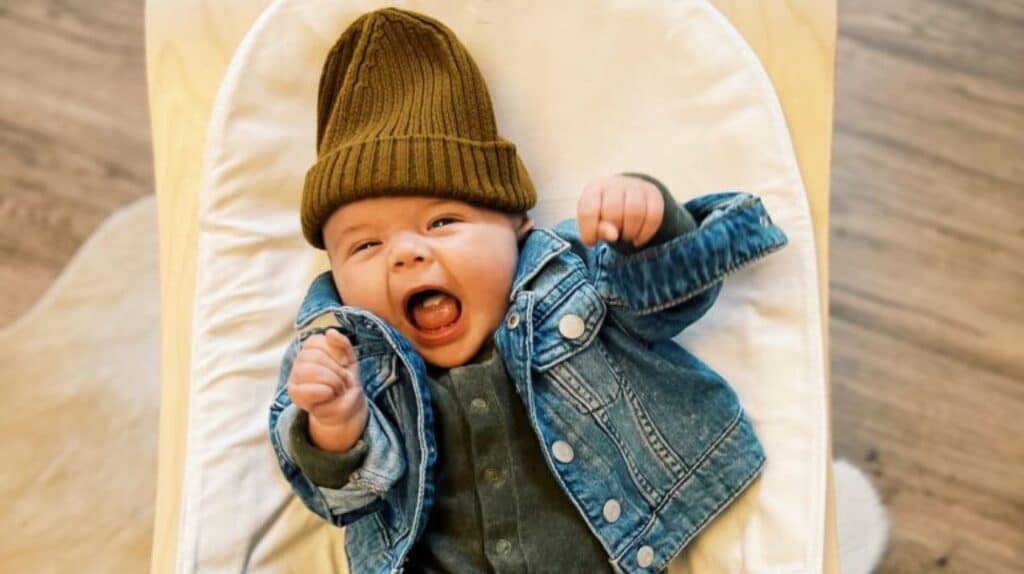 A baby wearing a denim jacket and a brown beanie, lying down with arms raised and mouth open as if excited or laughing.