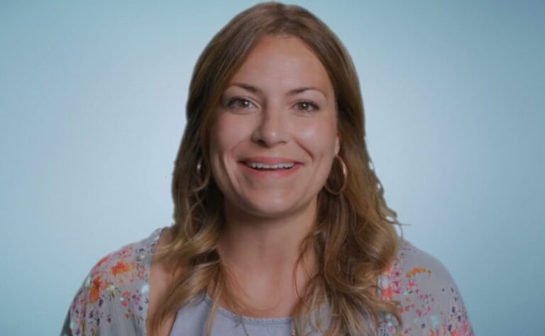 A woman with long brown hair, wearing a floral top, smiles against a light blue background.