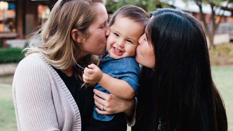 Two women kiss the cheeks of a smiling child they are holding outdoors, capturing a joyful moment together.