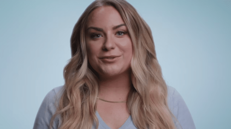 A person with long blonde hair smiles at the camera against a light blue background, wearing a light blue top and a necklace.