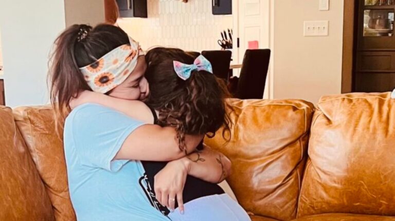 Two people with long hair hug each other warmly while sitting on a tan sofa in a cozy living room.