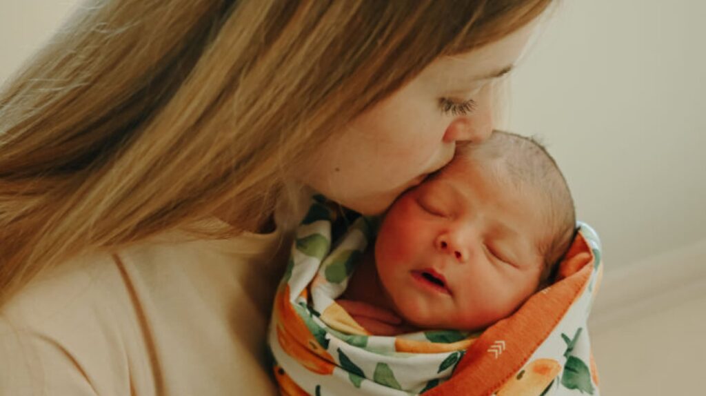 A woman kisses a sleeping newborn wrapped in a colorful blanket adorned with patterns of fruits and vegetables.