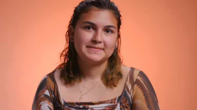 Person with shoulder-length hair wearing a patterned top, smiling softly against an orange background.