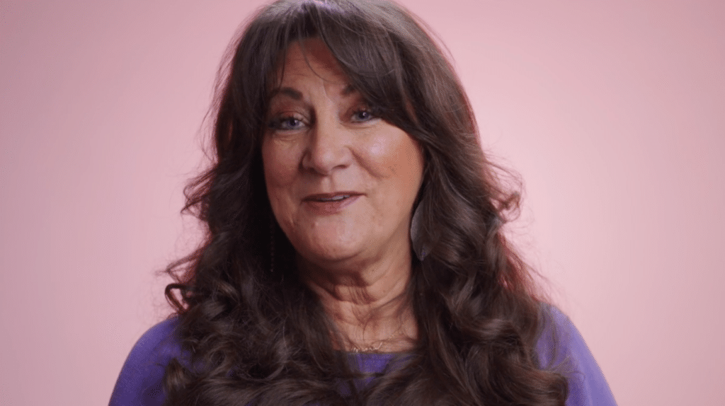 A woman with long, wavy brown hair smiles against a pink background, wearing a purple top.