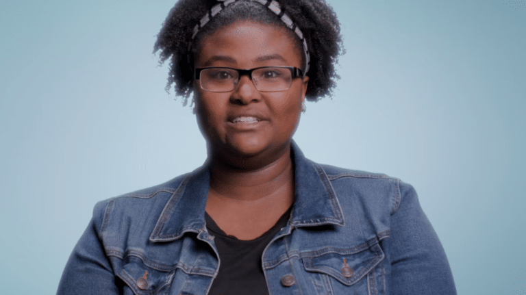 Person wearing glasses and a denim jacket against a light blue background, smiling slightly at the camera.