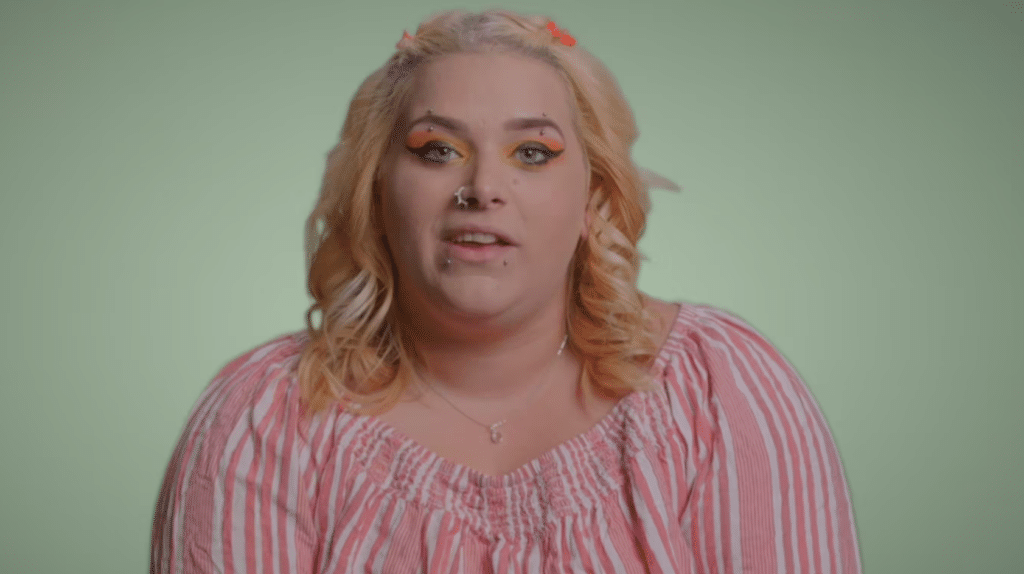 Person with blonde hair and colorful makeup, wearing a red-and-white striped top, against a plain green background.