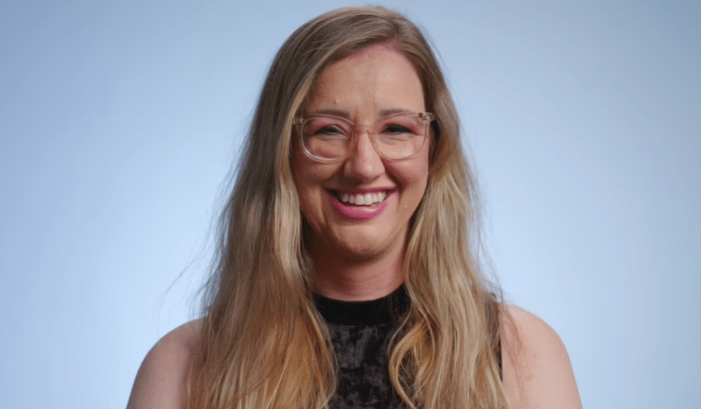 A smiling woman with long blonde hair and glasses wearing a dark top against a light blue background.