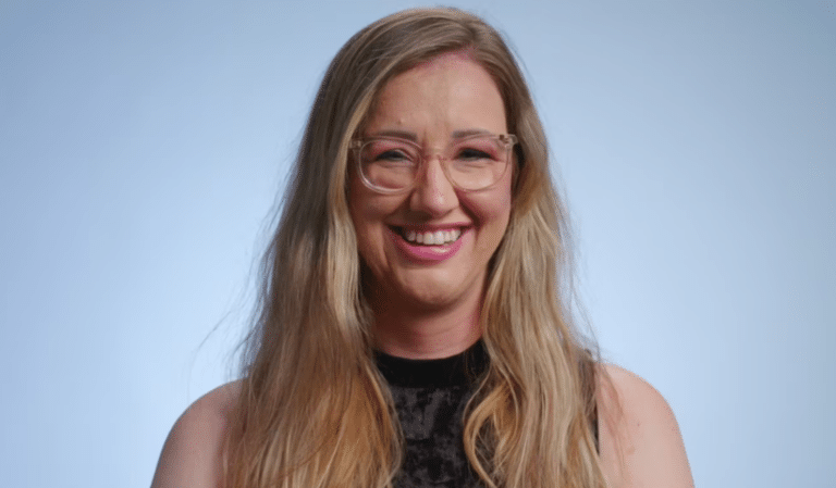 A smiling woman with long blonde hair and glasses wearing a dark top against a light blue background.