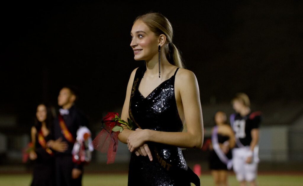 A young woman in a black sequin dress holds a rose and smiles at an outdoor night event, with people and lights blurred in the background.