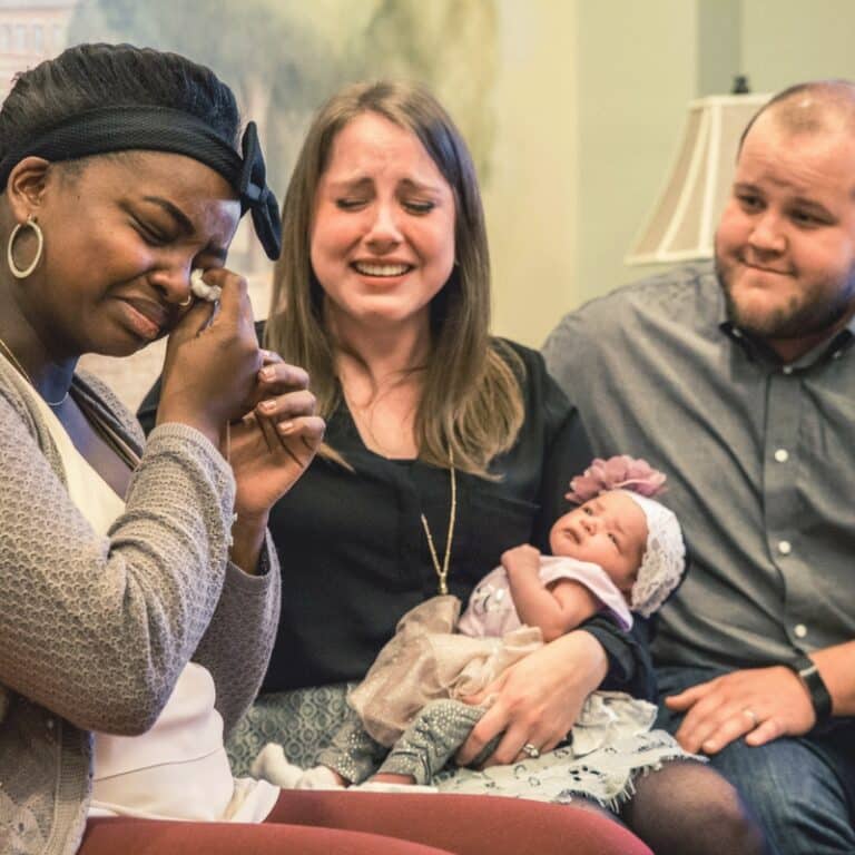 An emotional woman wiping tears, a couple holding a baby and smiling, all seated together; the setting appears to be indoors.