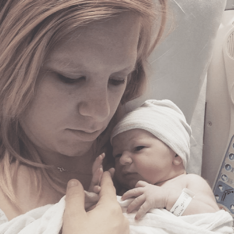 A woman cradles a newborn baby wrapped in a blanket and wearing a hat, both looking down.