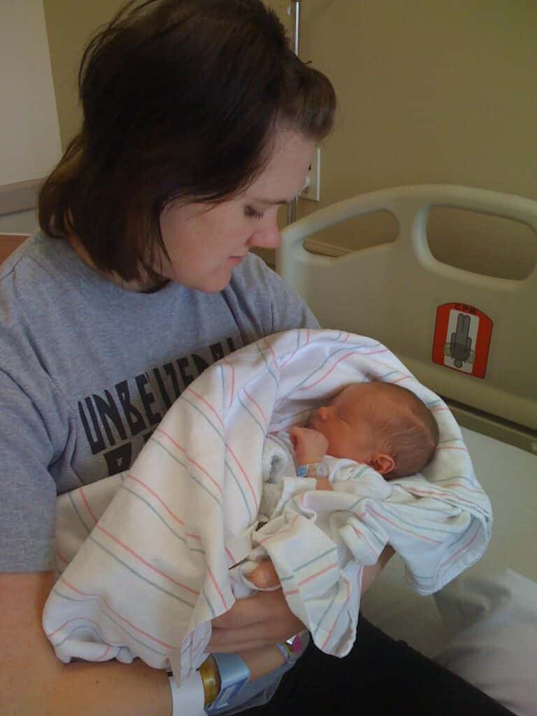 A woman in a gray shirt cradles a newborn wrapped in a striped blanket, sitting on a hospital bed.