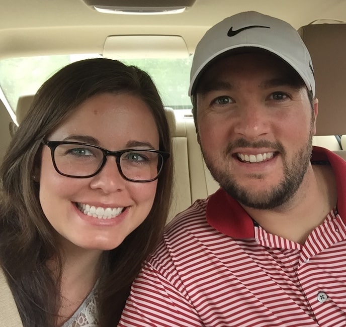 A smiling woman with glasses and a man in a striped shirt and cap pose for a selfie inside a car.