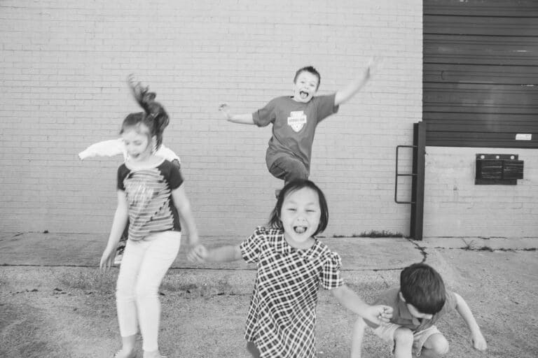 Four children are joyfully jumping and playing outdoors, with a brick wall in the background. The image is in black and white.