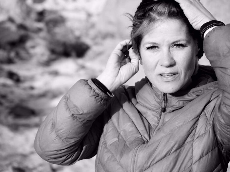 Black and white image of a person outdoors wearing a quilted jacket, looking at the camera and adjusting their hair with both hands.