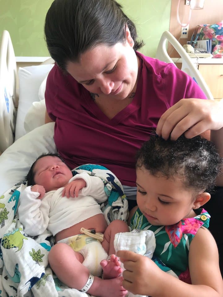 A woman in a hospital bed holds a newborn baby while a toddler sits nearby and touches their own hair.