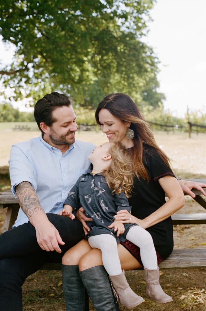 A family of three sits on a bench outdoors, smiling and laughing with each other on a sunny day.