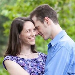 A couple in casual clothing smiling and embracing each other outdoors with greenery in the background.