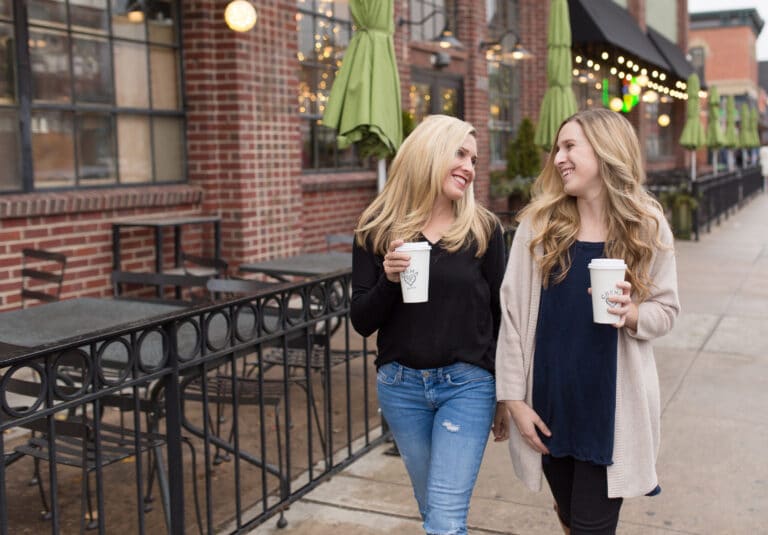 Two women walking outside, holding coffee cups, smiling and talking. They are in front of a brick cafe with green umbrellas.