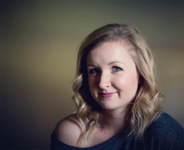 A woman with light hair and a slight smile, wearing a dark, off-the-shoulder top, sits in front of a soft, blurred background.