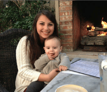 A smiling woman in a white sweater holds a baby in her lap. They sit at an outdoor table with a fireplace and greenery in the background.