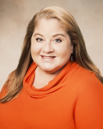 A woman with long blonde hair, wearing an orange top, smiles at the camera against a neutral background.