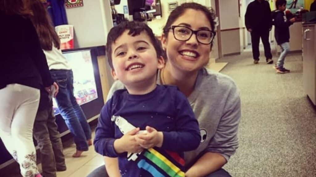 A woman with glasses and a child smile together indoors. The child holds a colorful toy, and people are seen in the background.