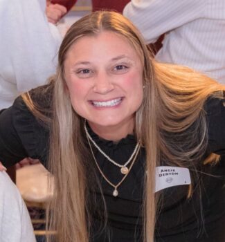 A smiling woman with long blonde hair wearing a black top and layered necklaces. She's wearing a name tag.