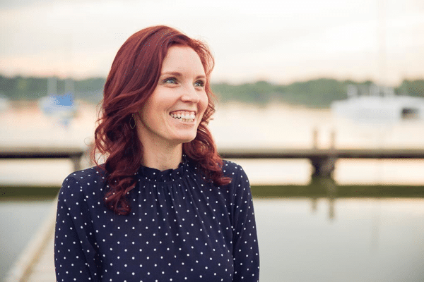 Smiling woman with red hair stands near a waterfront, wearing a navy blue polka dot blouse. Blurred boats and water in the background.