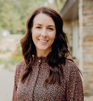 A woman with long dark hair and a floral blouse smiles while standing outdoors near a building.