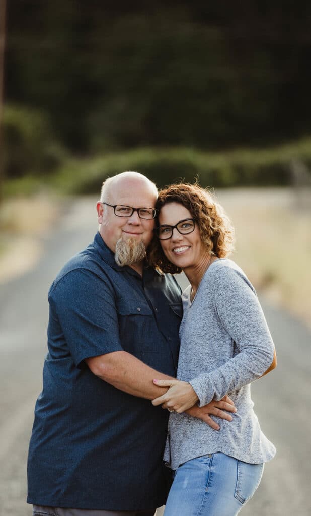 A couple wearing glasses, embracing and smiling on a gravel path with a blurred green landscape in the background.