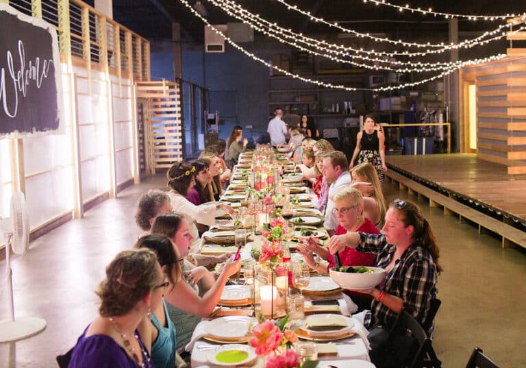 A long table filled with people enjoying a meal under string lights in an indoor venue with a warm, rustic atmosphere.