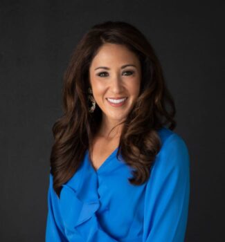 A woman with long, wavy brown hair smiles at the camera, wearing a bright blue blouse and drop earrings, against a dark background.