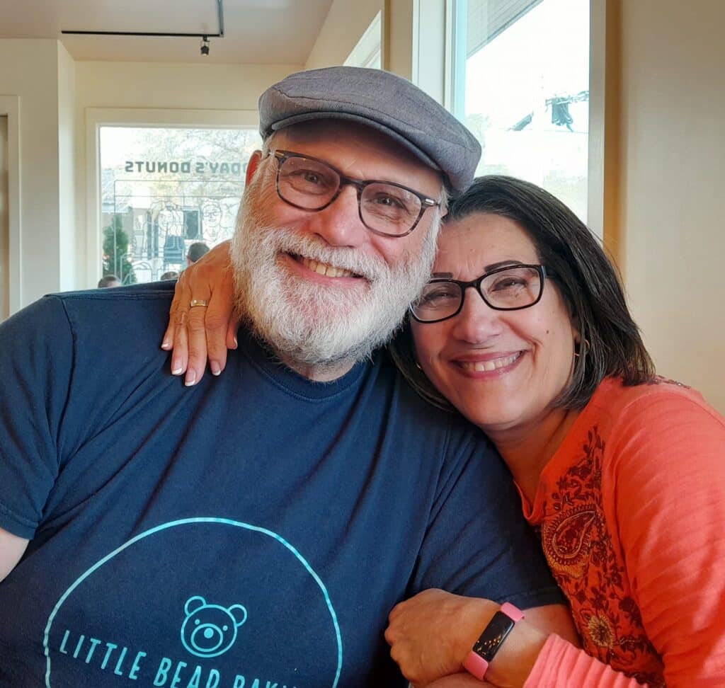 Two smiling people wearing glasses hugging; the man wears a hat and a "Little Bear Bakery" T-shirt, the woman wears a pink top.