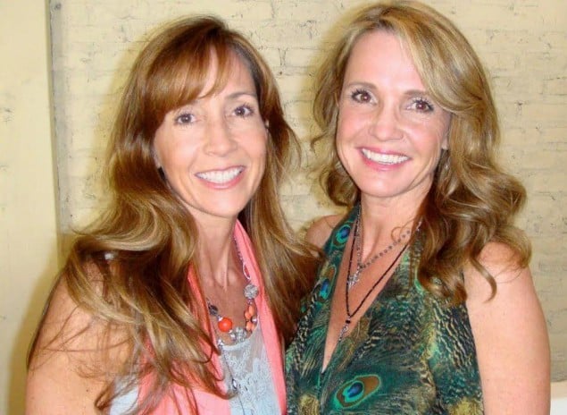 Two smiling women with long hair stand close together against a brick wall, wearing colorful sleeveless tops and necklaces.