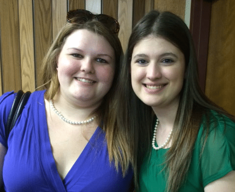 Two women smiling at the camera, one wearing a blue top and the other a green top. Both have long hair and are wearing pearl necklaces.