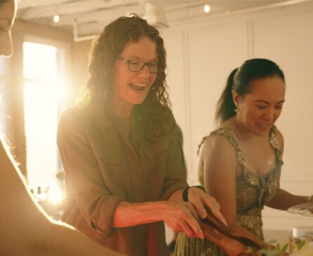 Three women smiling and serving food at a sunlit gathering.