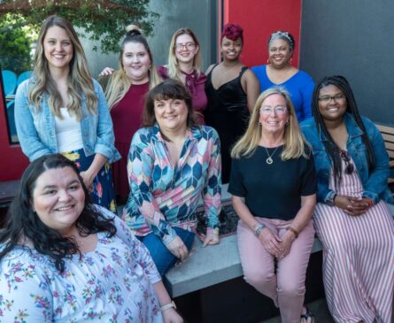 A group of nine women of diverse backgrounds posing together, smiling outdoors with a mix of casual and formal outfits.