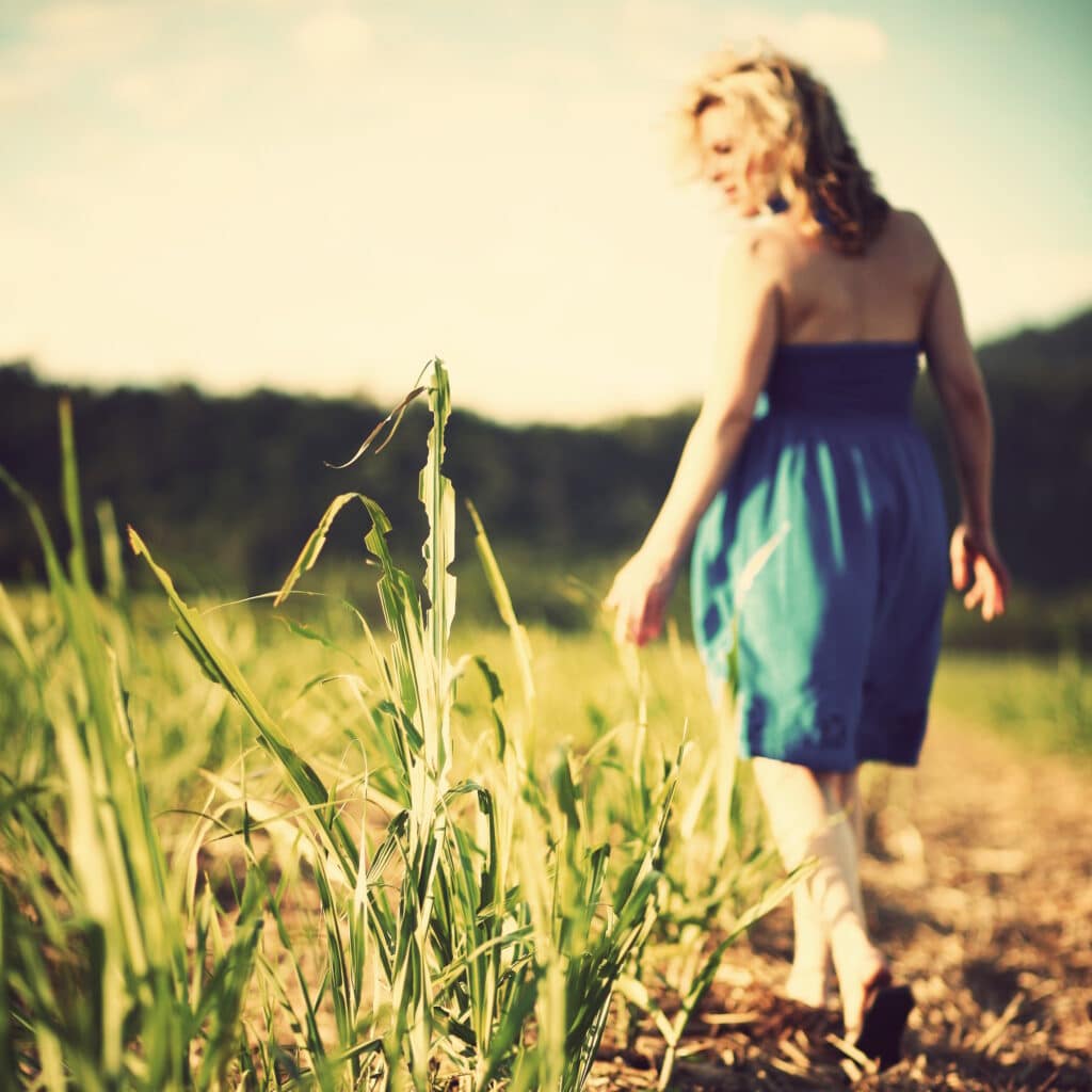 A woman in a blue dress walks barefoot through a field of tall grass on a sunny day, with a blurred background of greenery and hills.